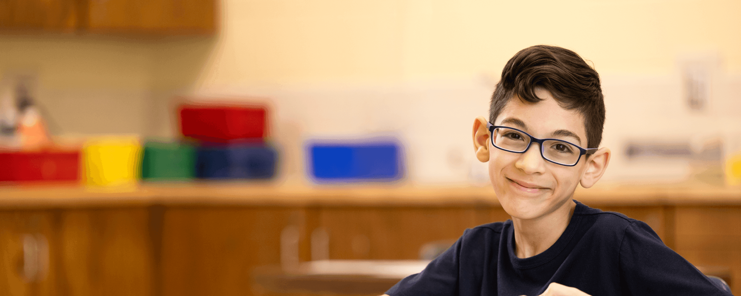 school aged boy with glasses sitting in a classroom and softly smiling at the camera